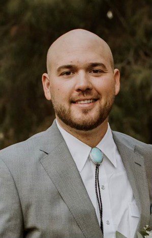 A smiling man in a gray suit with a turquoise bolo tie.
